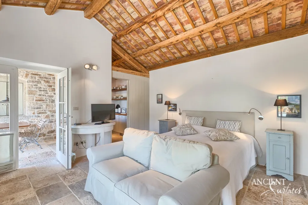 Stone-floored bedroom with a reclaimed timber-beam ceiling opening onto a limestone terrace